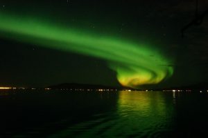 Green Northern Lights illuminating the night sky above a mountain, seen from a boat.